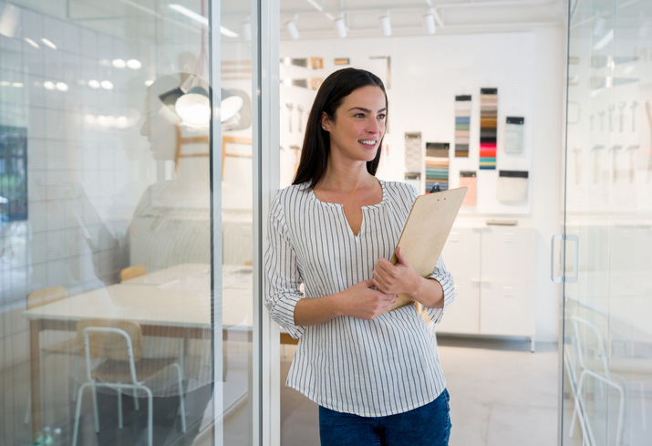 A confident female interior designer stands in a modern, light-filled office, holding a clipboard. Behind her, design materials and fabric samples are neatly arranged in a display area.